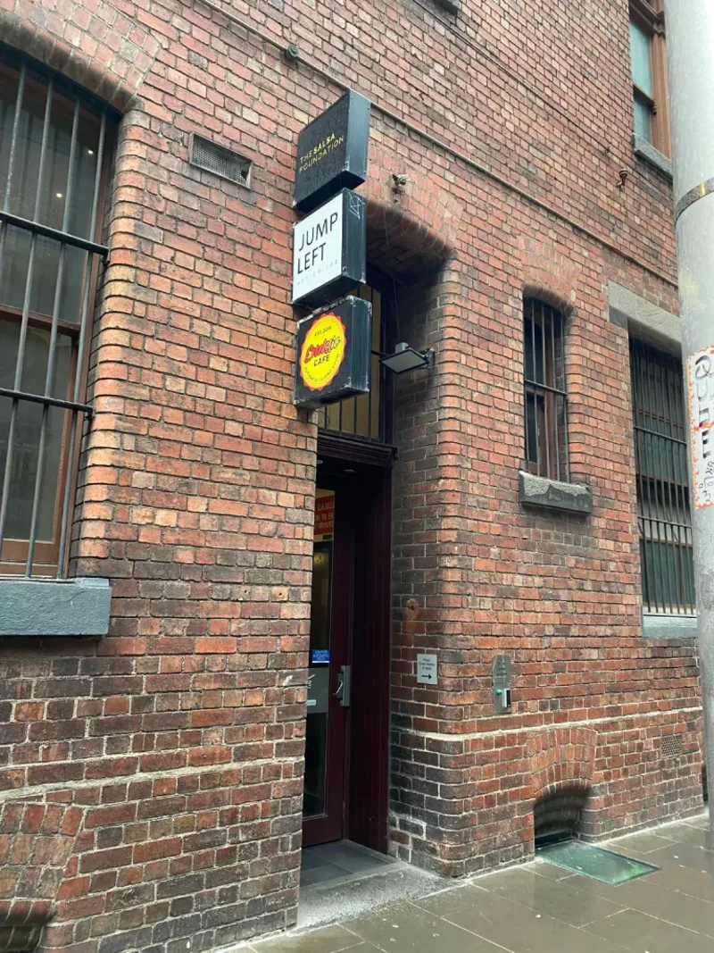 The Godfrey Street entrance: a recessed dark red door in a red brick wall, set back from the footpath, with three projecting signs stacked above it — "The Salsa Foundation", "Jump Left", and a round yellow cafe sign.